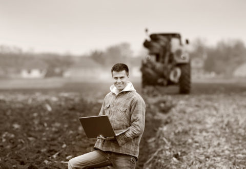 Young landowner with laptop supervising work on farmland, tractor in background, black and white image